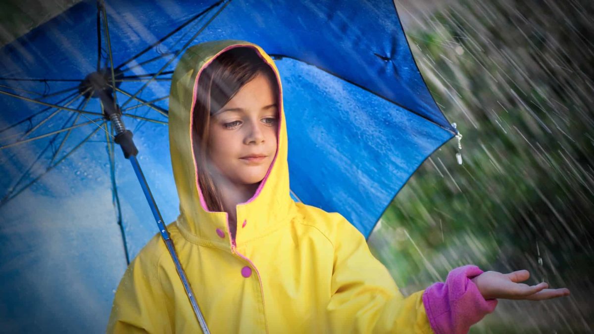 A young girl in a yellow raincoat holds a big blue umbrella in the pouring rain, frowning while the rain falls onto her held out hand.