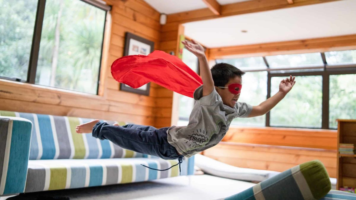 Child with superhero mask and cape flies after jumping on sofa