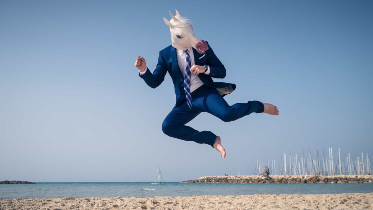 A man in a horse head mask and suit jumps for joy on a beach.