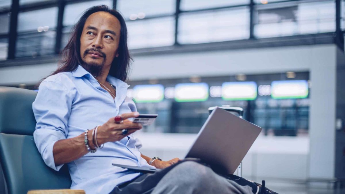 A man sits in the airport terminal with a laptop and credit card, ready to make a travel booking.