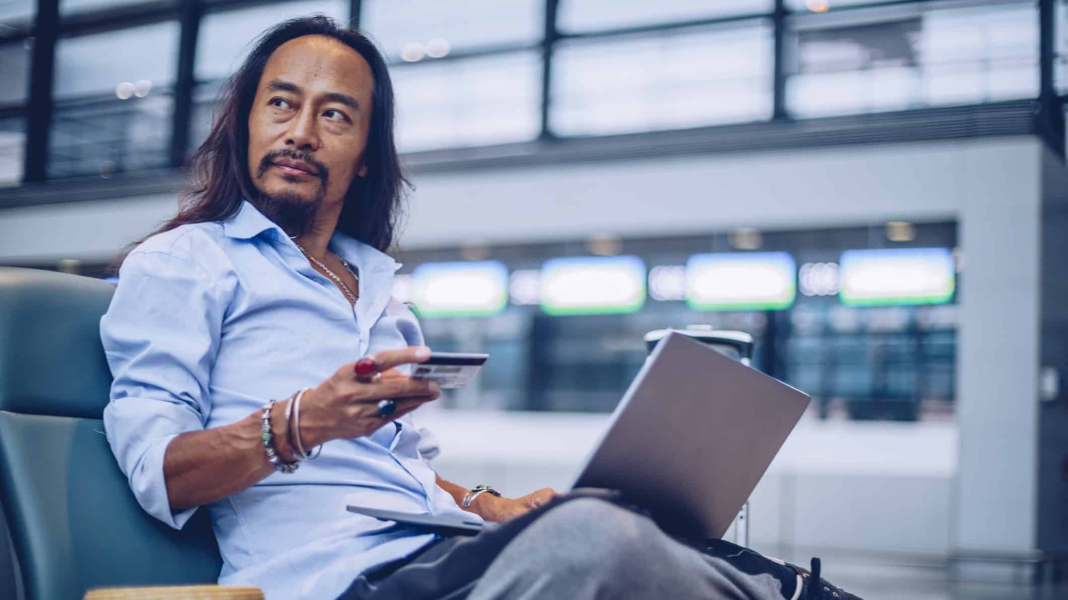 A man sits in the airport terminal with a laptop and credit card, ready to make a travel booking on the Flight Centre