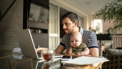 A man holds his baby on his lap at the dining room table while he looks at his laptop screen earnestly.