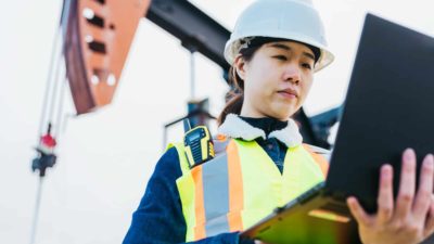 Female miner in hard hat and safety vest on laptop with mining drill in background.