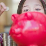 Young girl peeps over the top of her red piggy bank, ready to put coins in it.