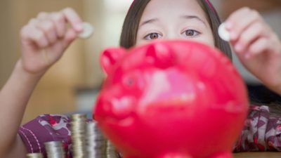 Young girl peeps over the top of her red piggy bank, ready to put coins in it.