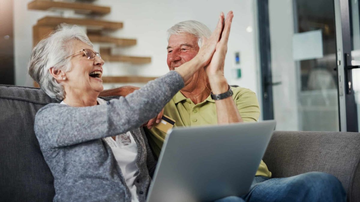 An older woman high fives an older man with big smiles after seeing good news on their laptop regarding their ASX tech shares