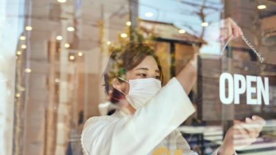 A woman wearing a face mask turns the open sign in the glass door of the shop.