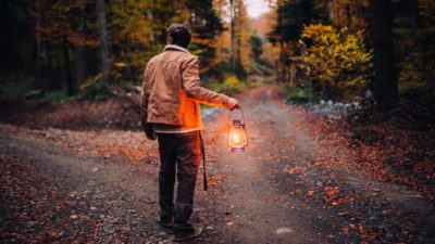 A man faces a fork in the path in the bush before being plunged into the night's darkness holding only a gas lantern.