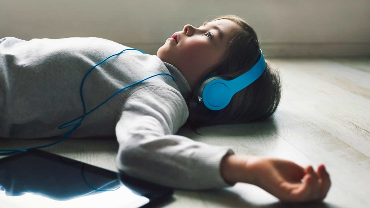 A kid lies on the floor and stares up at the ceiling with headphones over his ears and a tablet next to him.