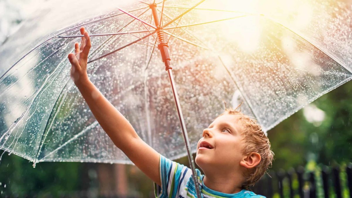 A young boy reaches up to touch the raindrops on his umbrella, as the sun comes out in the sky behind him.