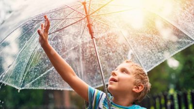 A young boy reaches up to touch the raindrops on his umbrella, as the sun comes out in the sky behind him.