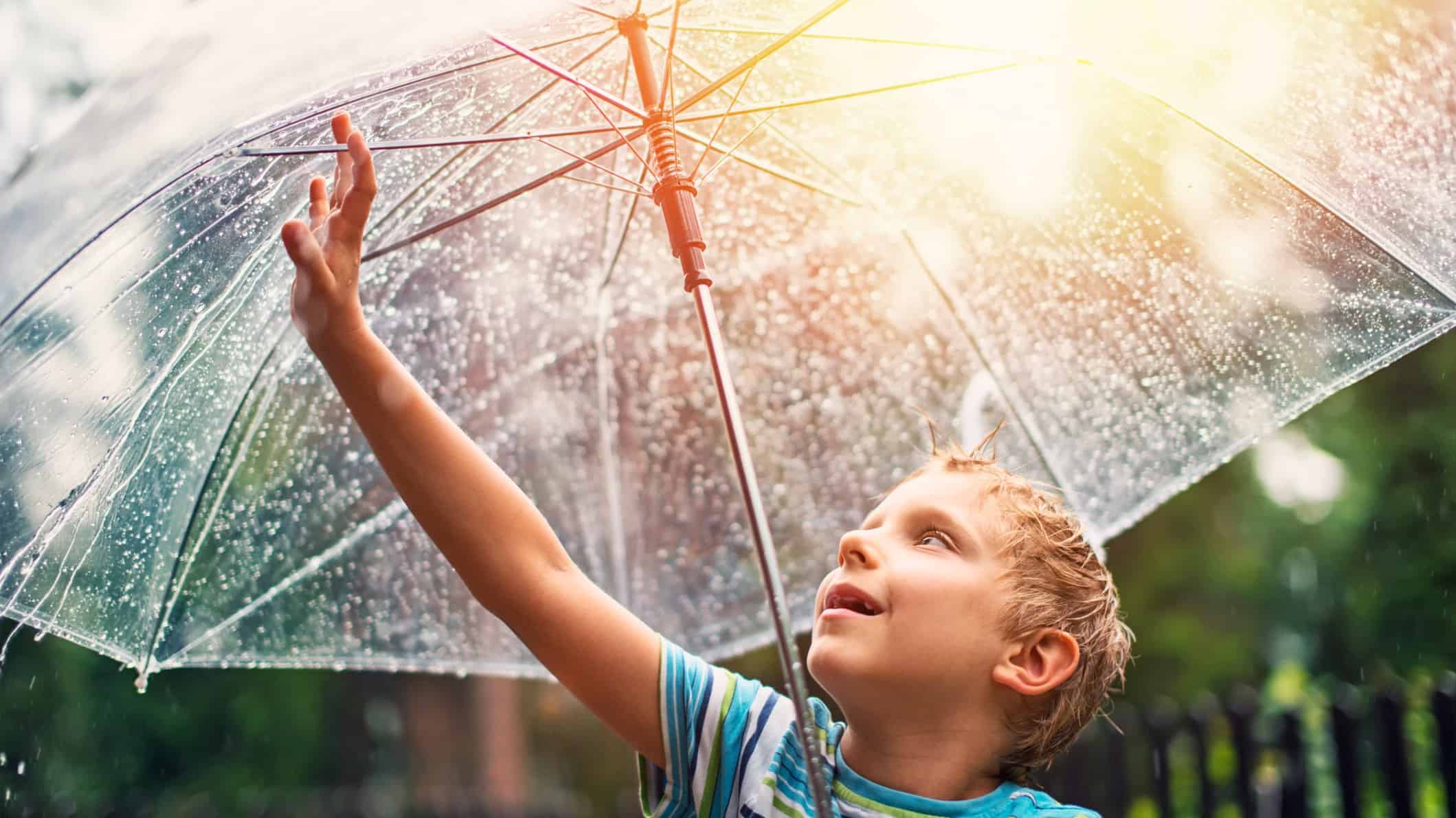 A young boy reaches up to touch the raindrops on his umbrella, as the sun comes out in the sky behind him representing the rising Fortescue share price due to heavy rain in Brazil recently