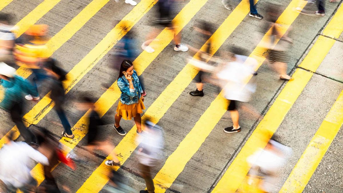 A woman walks slowly across a yellow pedestrian crossing while people around her walk so fast they are blurred.