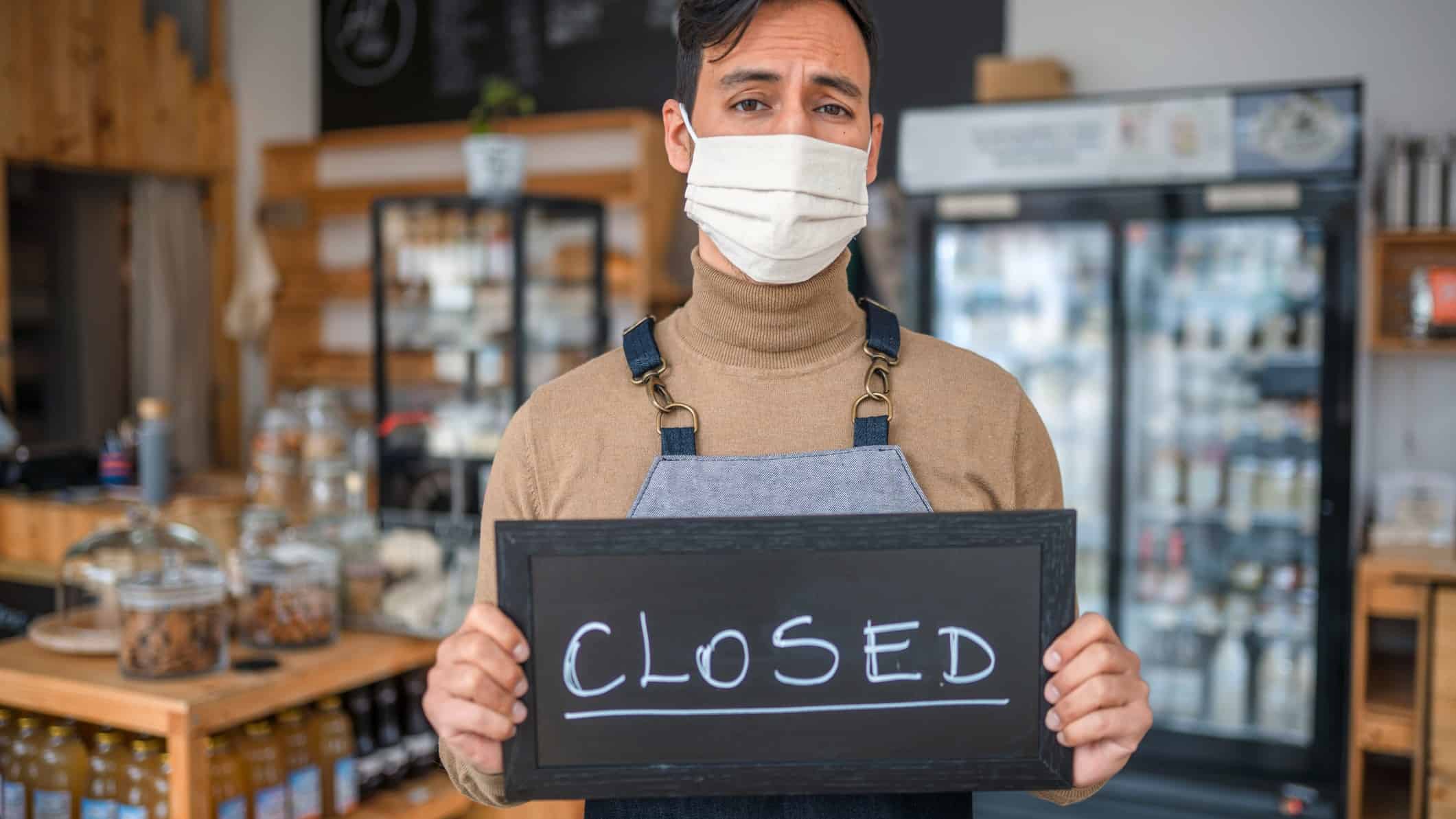 A masked shopkeeper holds a closed sign in his empty store.