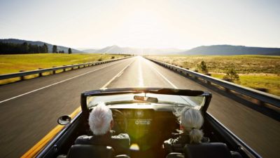 An older couple driving in a convertible on a freeway.