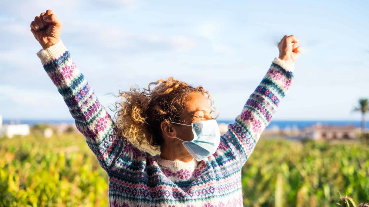 A happy masked woman is vaccinated, COVID-free and winning with both hands in the air.