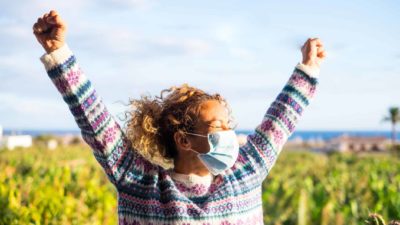 A happy masked woman is vaccinated, COVID-free and winning with both hands in the air.