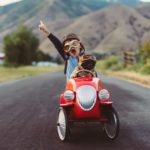 a child in a billy cart style car holds a hand in the air as he drives ahead on an open road.