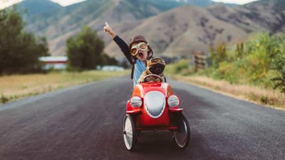 a child in a billy cart style car holds a hand in the air as he drives ahead on an open road.