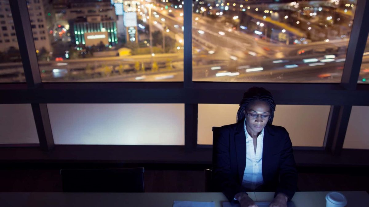 Woman sits at her desk working at night, while traffic flows on a busy freeway out the window behind her.