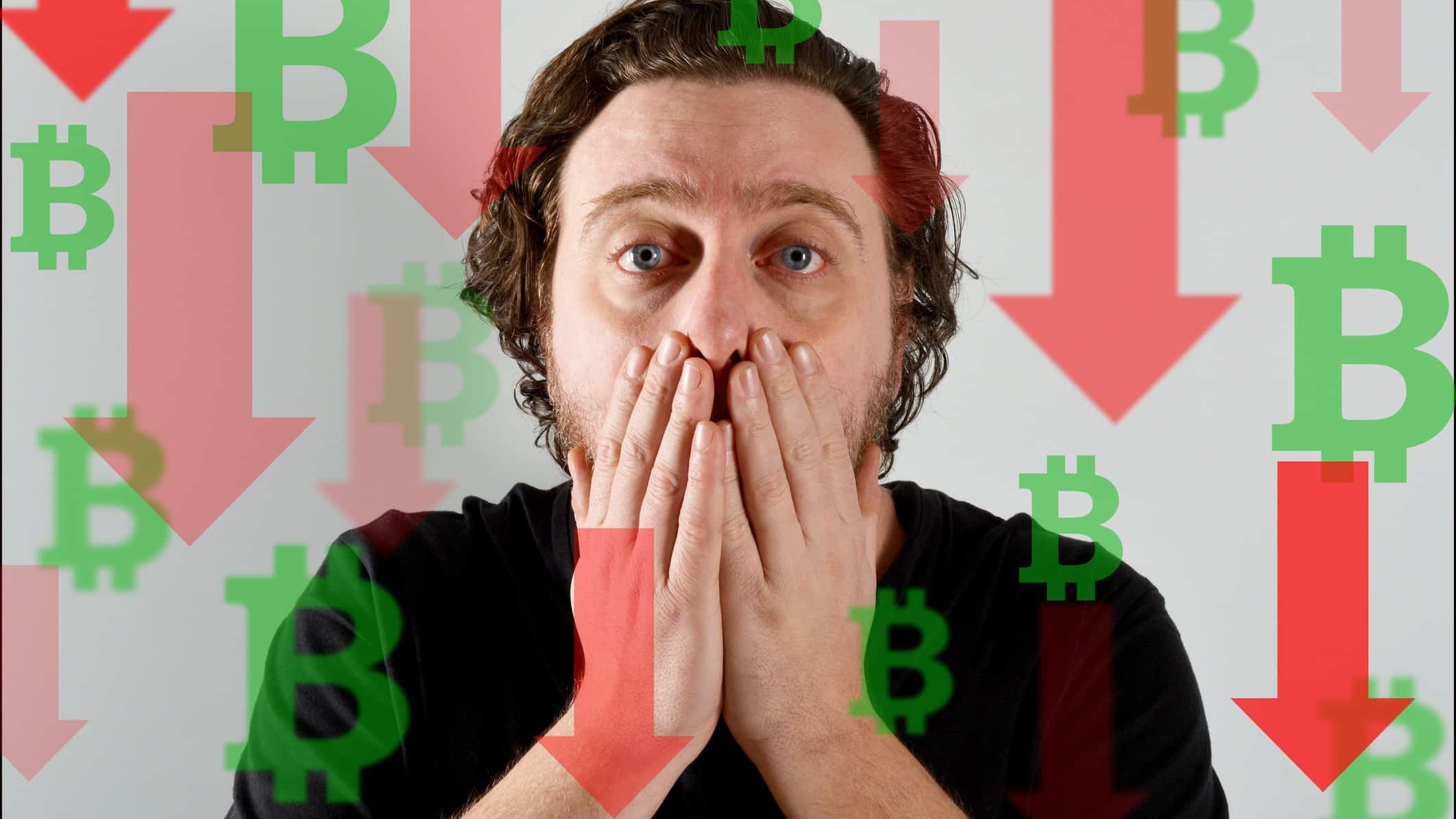 A bitcoin trader looks afraid and holds his hands to his mouth among graphics of red arrows pointing down