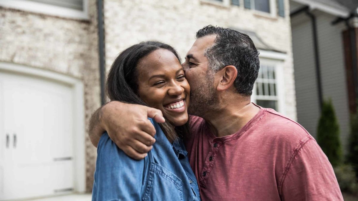 Man puts arm around woman and kisses her cheek outside their new home