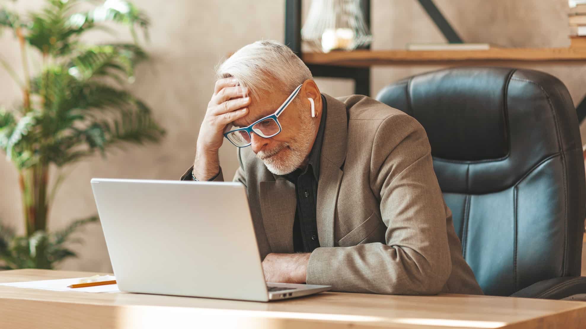 Disappointed elderly man with regret sits with head in hand at computer