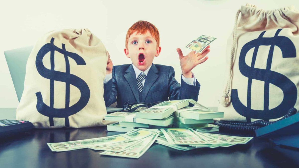 A boy hold money and dressed in business suit next to money bags on a desk, indicating a dividends windfall