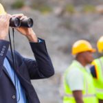 Engineer with hard hat looks through binoculars at work site or mine as two workers look on