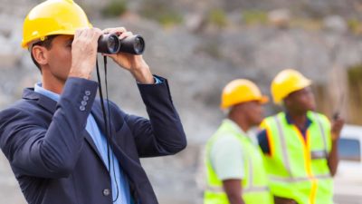 Pilbara Minerals engineer with hard hat looks through binoculars at work site or mine as two workers look on