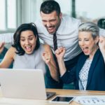 Three excited business people cheer around a laptop in the office