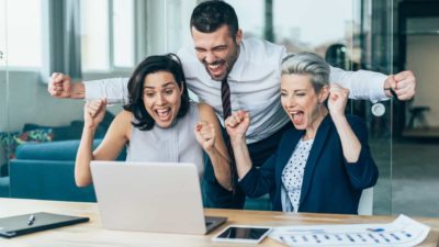 Three excited business people cheer around a laptop in the office