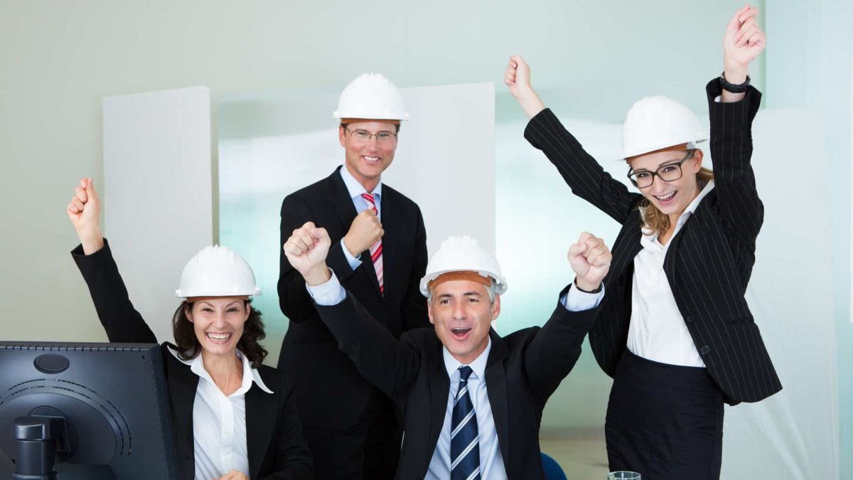 Four people in business suits and white hard hats sit in front of desk and cheer