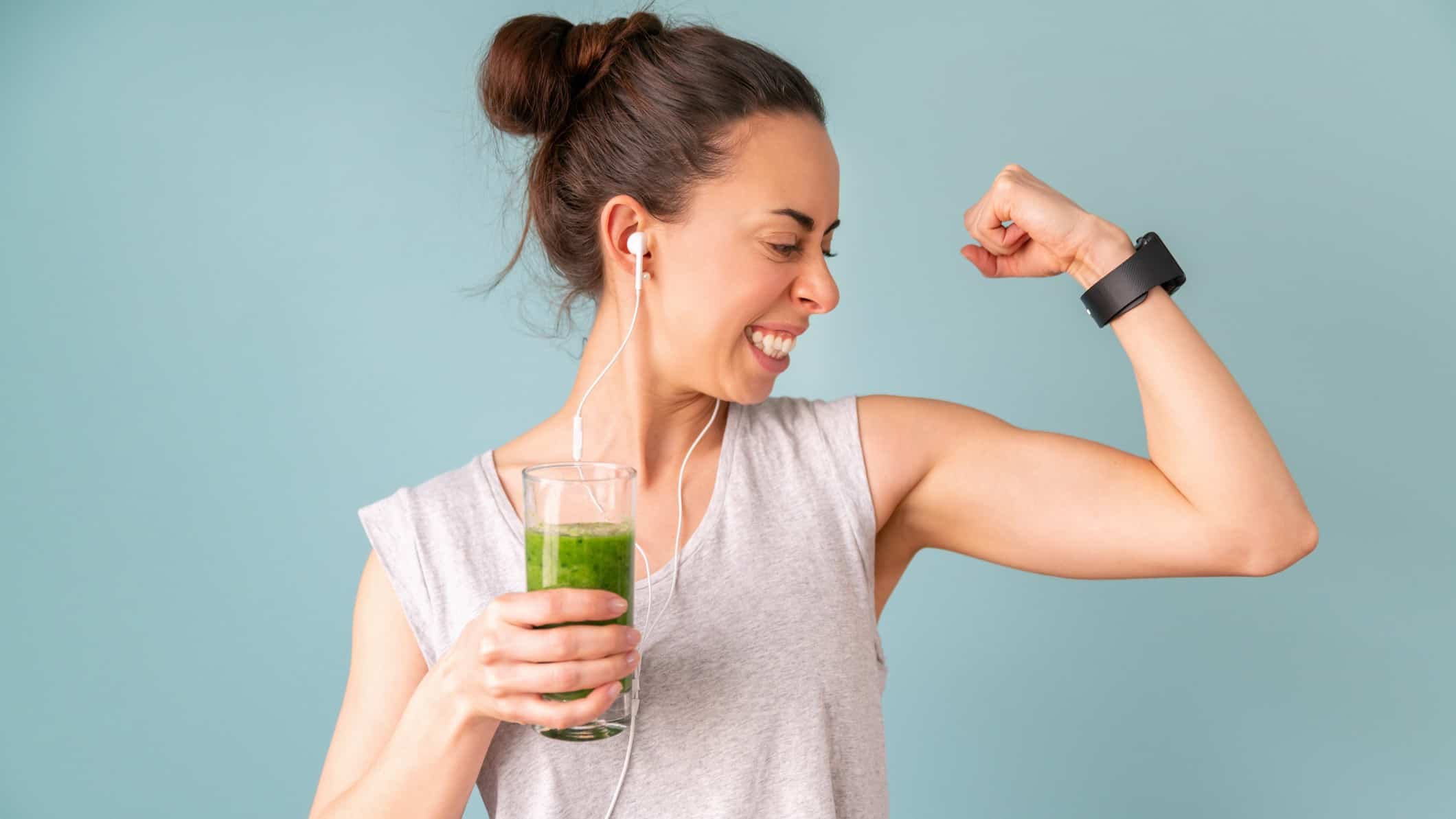 A woman in workout gear flexes her muscles while holding a juice