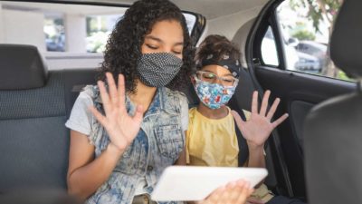 Two children in car wearing covid masks and playing on laptop