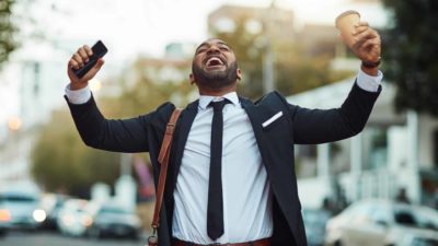 Man puts hands in the air and cheers with head back while holding phone and coffee