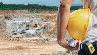 A miner holding a hard hat stands in the foreground of an open cut mine