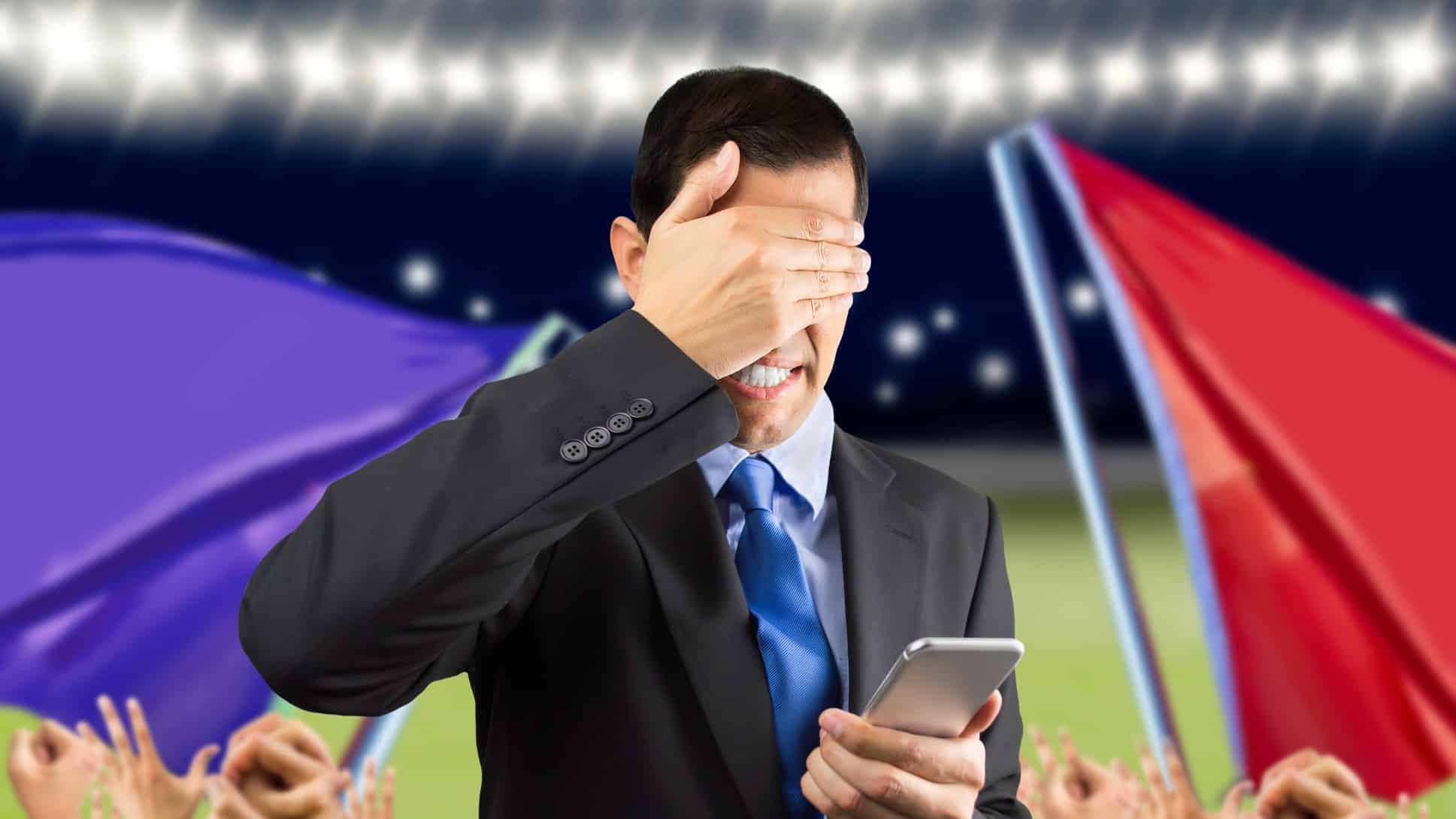 Man puts hand over face as he loses online bet at stadium with flags behind him