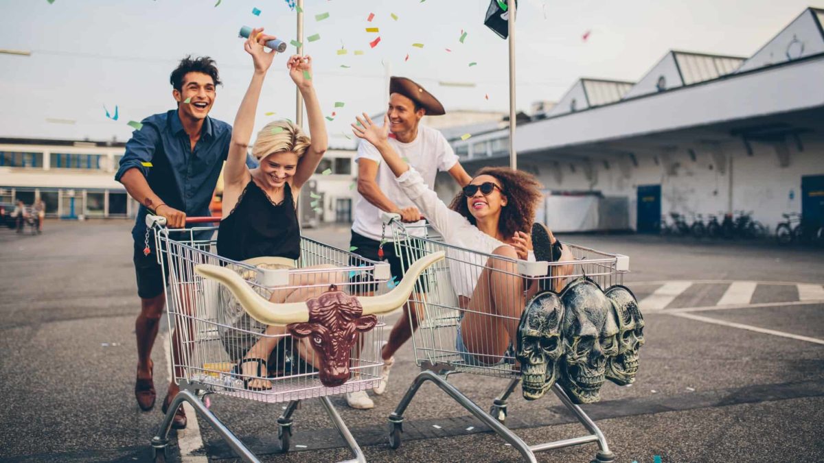 Two couples race each other in supermarket trollies, having a great time, smiling and laughing.
