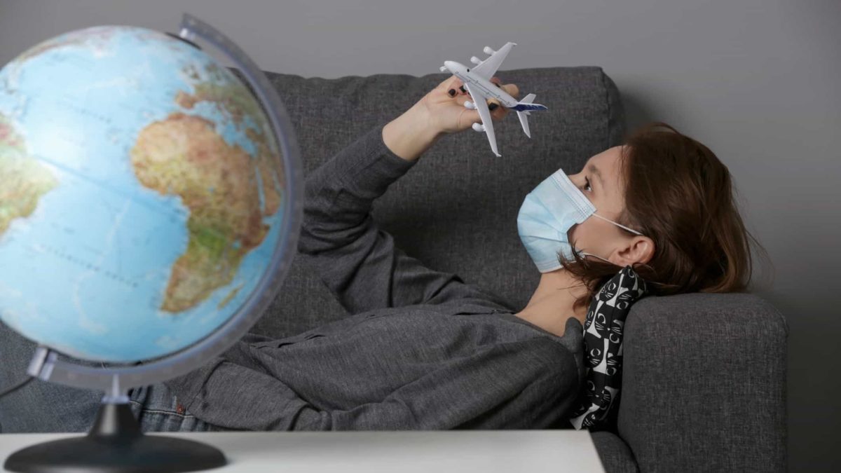 A woman wearing a facemask slumps on a couch next to a globe of the world, indicating COVID travel restrictions in play