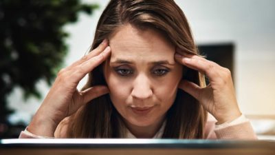 Woman sits at computer in a quandary with hands at side of head