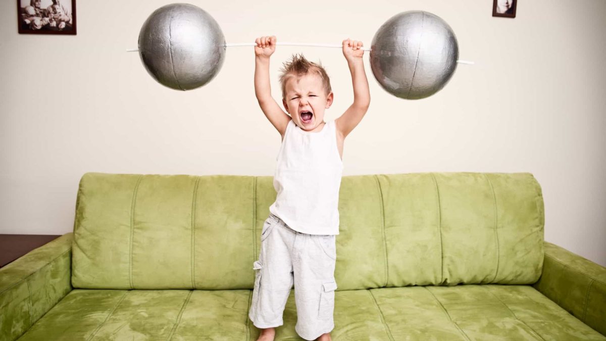 A young boy lifts a barbell over his head while standing on a couch.