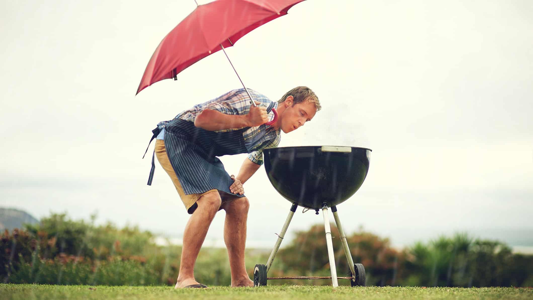 A man holds an umbrella and tries to ignite his bbq while rain pours down.