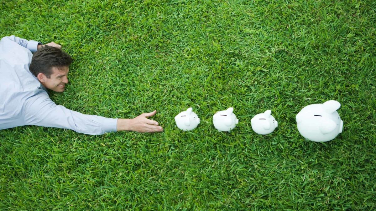 A man sprawls on the grass reaching out to touch four piggy banks, lined up in a row.