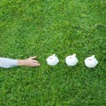 A man sprawls on the grass reaching out to touch four piggy banks, lined up in a row.