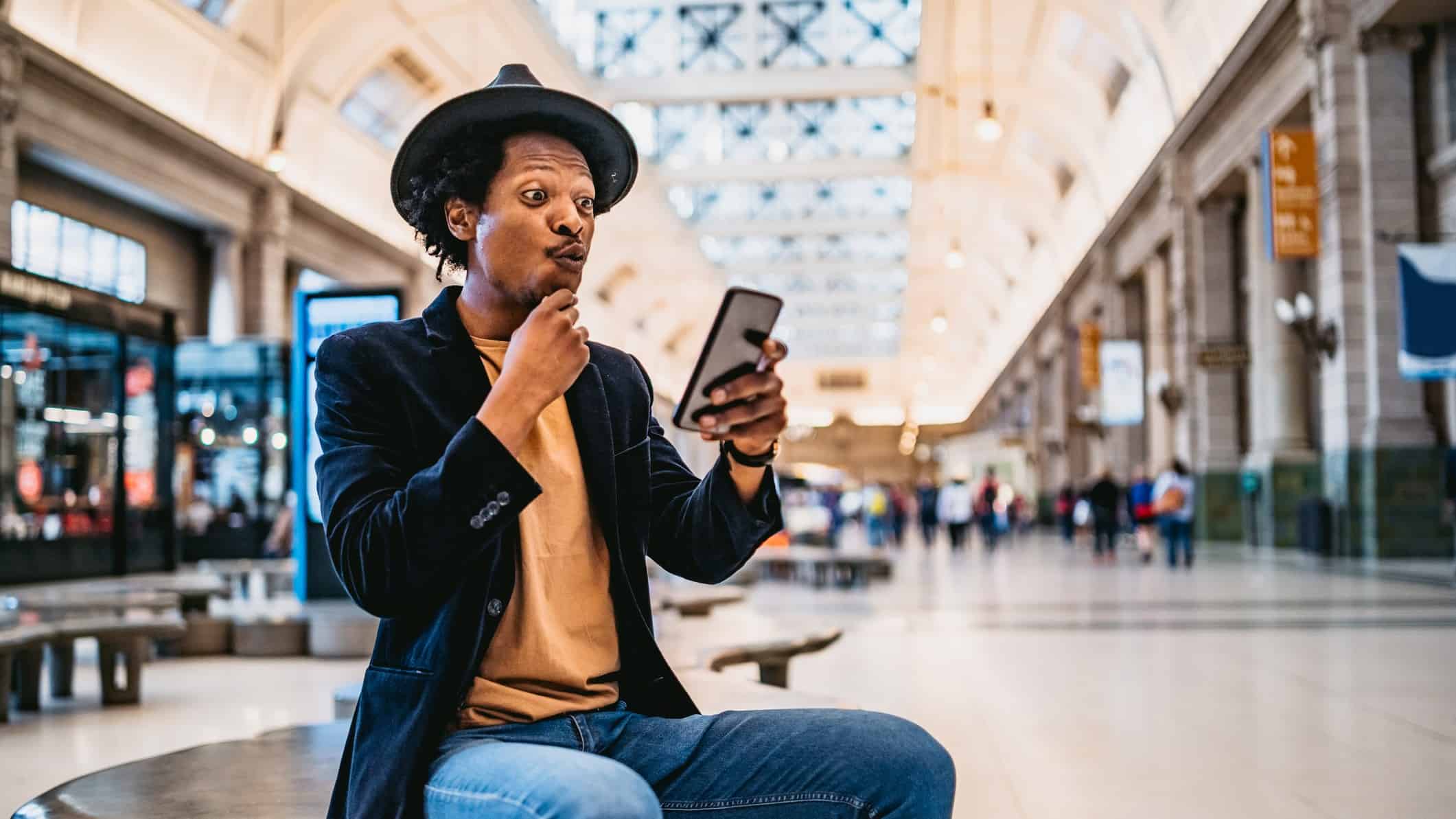 A man in trendy clothing sits on a bench in a shopping mall looking at his phone with interest and a surprised look on his face.