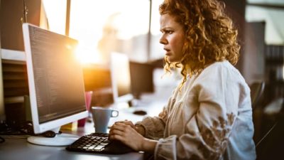 A woman sees bad news on her computer screen while the morning sun rises through the window behind her.