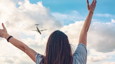 A woman looks up at a plane flying in the sky with arms outstretched as the Flight Centre share price surges