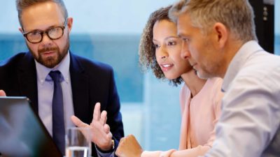 A man in a suit looks serious while discussing business dealings with a couple as they sit around a computer at a desk in a bank home lending scenario.