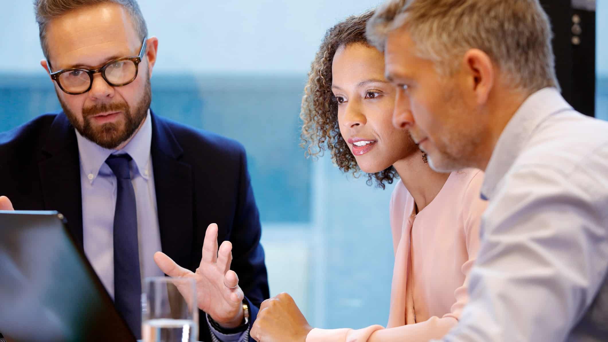 a man in a suit looks serious while discussing business dealings with a couple as they sit around a computer at a desk in a bank home lending scenario.
